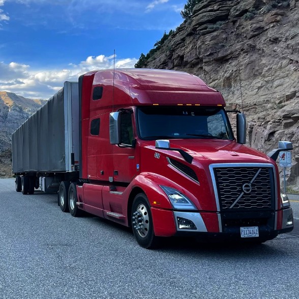 Red Volvo tractor pulling a Conestoga rolling-tarp trailer through a mountain pass.